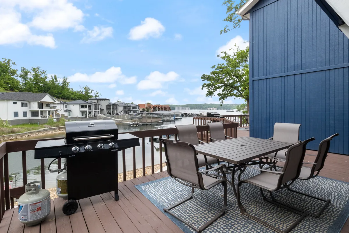 An exterior dining area overlooking the water, with a grill, large dining table, lush greenery, and expansive water views—ideal for outdoor meals, socializing, and soaking up the lakeside vibe.