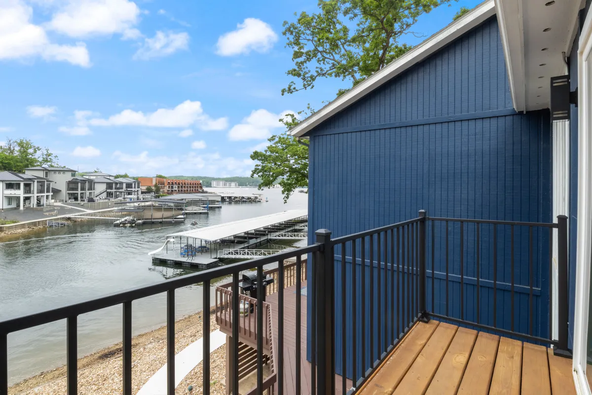 A tranquil waterfront view from a balcony, featuring a blue exterior railing and house, with expansive views of the lake and boats passing by. The calm water and greenery create a sense of relaxation and escape.