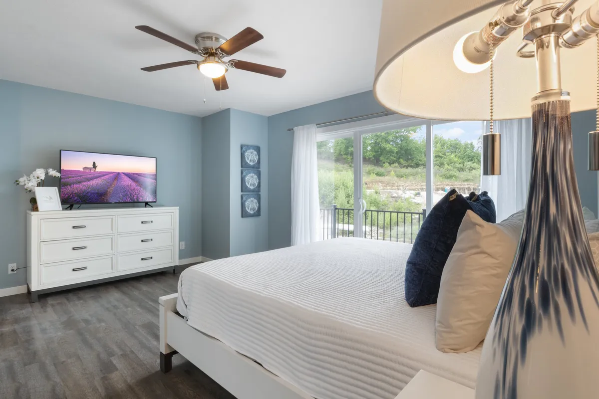 A modern bedroom with a cozy bed topped with decorative pillows, a flat-screen television, a large window allowing in morning light, and a dresser for personal storage. The decor is simple yet elegant, emphasizing comfort and personal sanctuary.