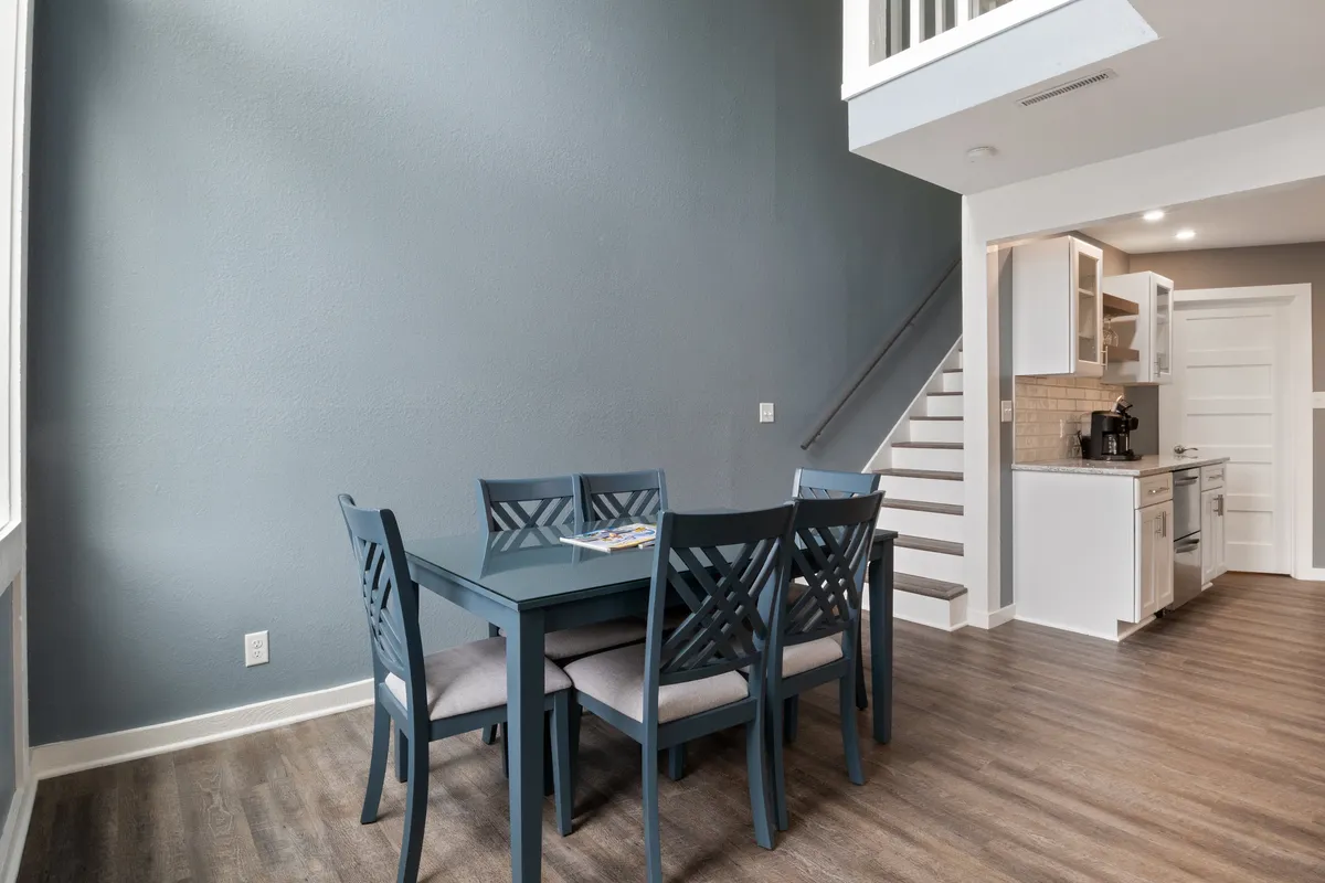 A modern dining area with a table, staircase, and kitchen, set in a bright, open interior. The table is ready for group meals, game nights, or celebratory toasts among friends.