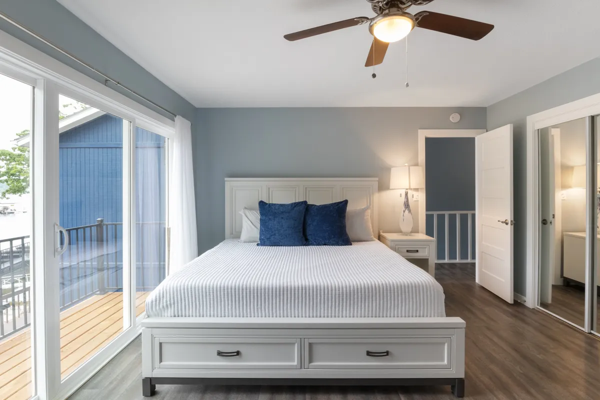 This image features a spacious bedroom with a comfortable bed adorned with blue accent pillows, large windows that let in abundant natural light, a ceiling fan for added comfort, and a nightstand with a lamp for evening relaxation. The serene atmosphere is perfect for restful nights and calm mornings.