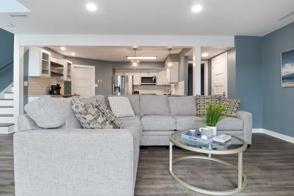 This image presents a large gray sectional sofa and a glass coffee table, with a view into the modern kitchen area in the background. The open-plan design and spacious seating highlight an inviting area for groups to gather, share stories, or prepare for a night out.