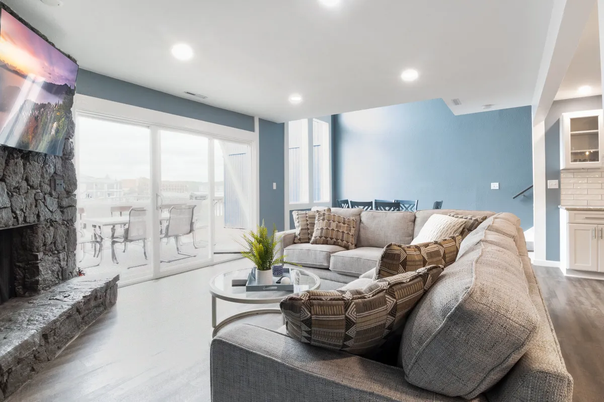 An inviting living area with a large sectional sofa, stone fireplace, television, and a dining area visible through glass doors. The room is bathed in natural light, with cozy, welcoming furnishings and clear integration of dining and relaxation spaces.