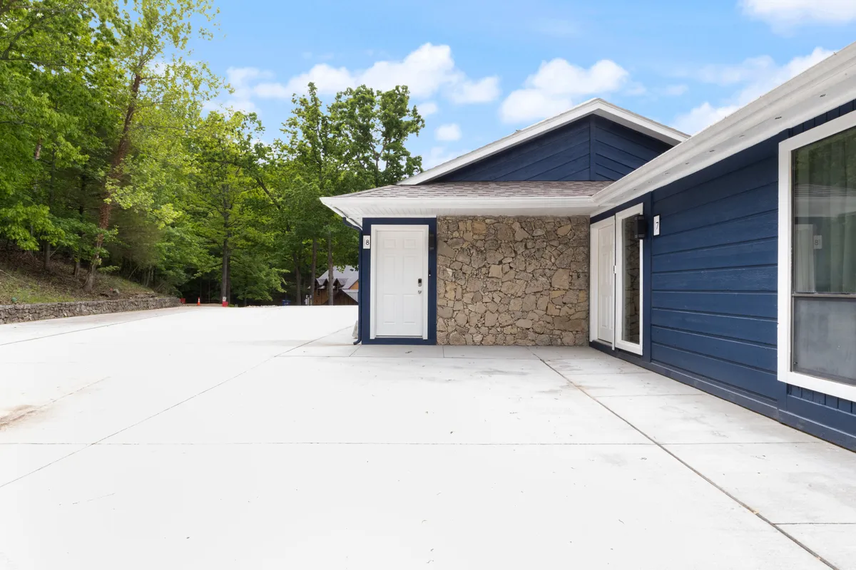 This image features the exterior of the property, showcasing blue wooden siding, a natural stone wall, and a welcoming front door. The sunny outdoor space hints at areas for gathering, relaxing, and enjoying the fresh air, with ample room for family activities.
