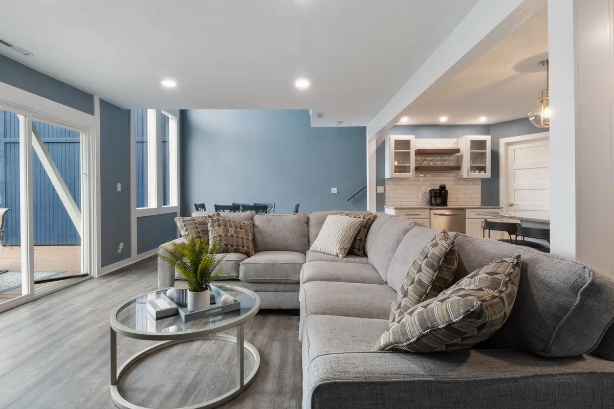 A spacious open-plan interior featuring a large sectional sofa, glass coffee table, and a modern kitchen area with large windows letting in natural light. The space is designed for socializing and relaxation, with clear sightlines across living, dining, and kitchen zones.
